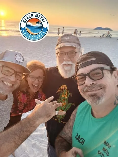 Four people taking a selfie on a beach during sunset, with the Beach Metal Detectives logo. They are smiling and appear happy, with the ocean and some distant people and islands in the background.