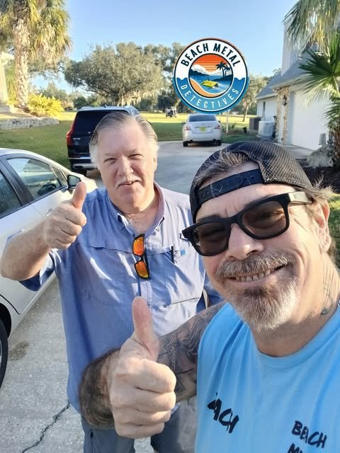 Two men smiling and giving thumbs up in front of a car, with a beach-themed Detective sign in the background. One man is wearing sunglasses and a baseball cap, while the other is wearing a blue shirt and sunglasses hanging on his shirt collar.