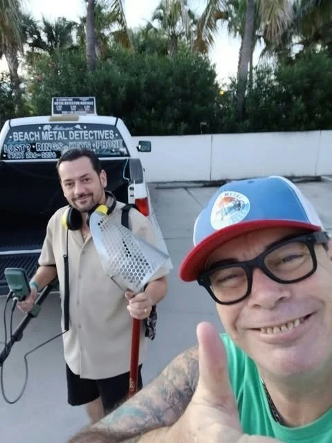 Two smiling men taking a selfie in a parking lot with palm trees in the background. One man is wearing a trucker hat, glasses, and a green shirt, giving a thumbs up. The other man is holding a metal detector and has headphones around his neck. Behind them is a black truck with a sign on top that reads 'Beach Metal Detectives' and includes contact information.