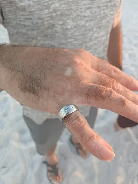 A close-up of a person's hand with a silver ring on the ring finger, showing some white residue or chalk, with sandy ground in the background.