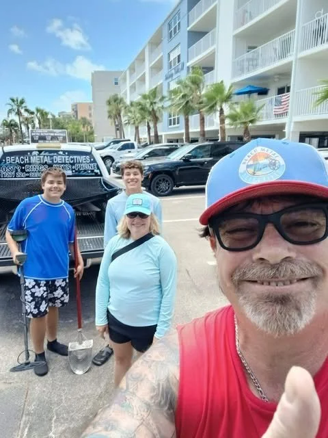 A man taking a selfie with three children in a parking lot near a beach, with palm trees and multi-story buildings in the background.