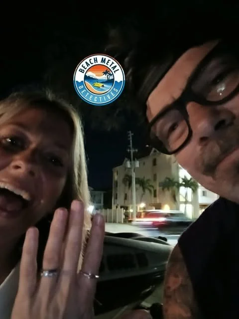 A woman and a man taking a selfie in a vehicle at night, with a Beach Metal Detectives sticker and houses with palm trees in the background.