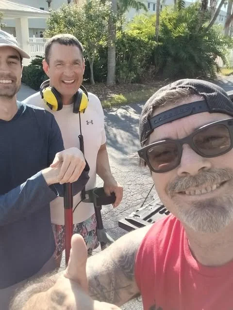 Three smiling men outdoors taking a selfie, one giving a thumbs up, with trees and houses in the background.