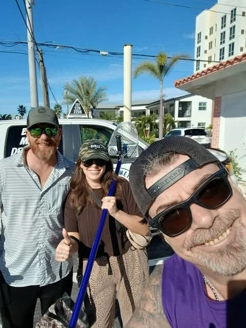 Three people smiling outdoors, taking a selfie. One man in the foreground with a beard, sunglasses, and a hat, wearing a purple shirt. A woman in the middle holding a lacrosse stick, wearing sunglasses and a baseball cap. Another man on the left wearing sunglasses, a gray hat, and a striped shirt. There are palm trees and a white building in the background.
