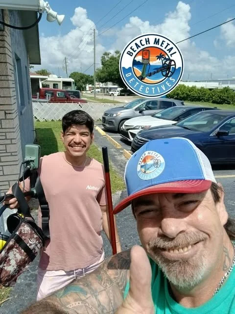 Two smiling men standing outside near parked cars, holding fishing gear, with a circular sign that reads "Beach Metal Detectives" in the background.