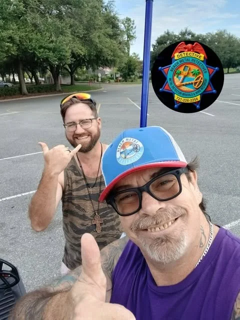 Two smiling men taking a selfie outdoors on a parking lot with trees in the background. One man is wearing glasses, a blue and red baseball cap, and a purple shirt. The other man is wearing glasses, a necklace, and a sleeveless shirt. A detective badge sticker is edited above them.