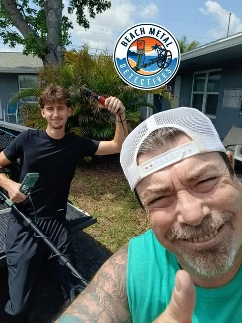 Two men smiling outdoors, one in the foreground wearing a white cap and teal shirt giving a thumbs up, the other in the background holding a fishing lure, with a logo reading 'Beach Metal Detectives' featuring a beach, sunset, and metal detector.