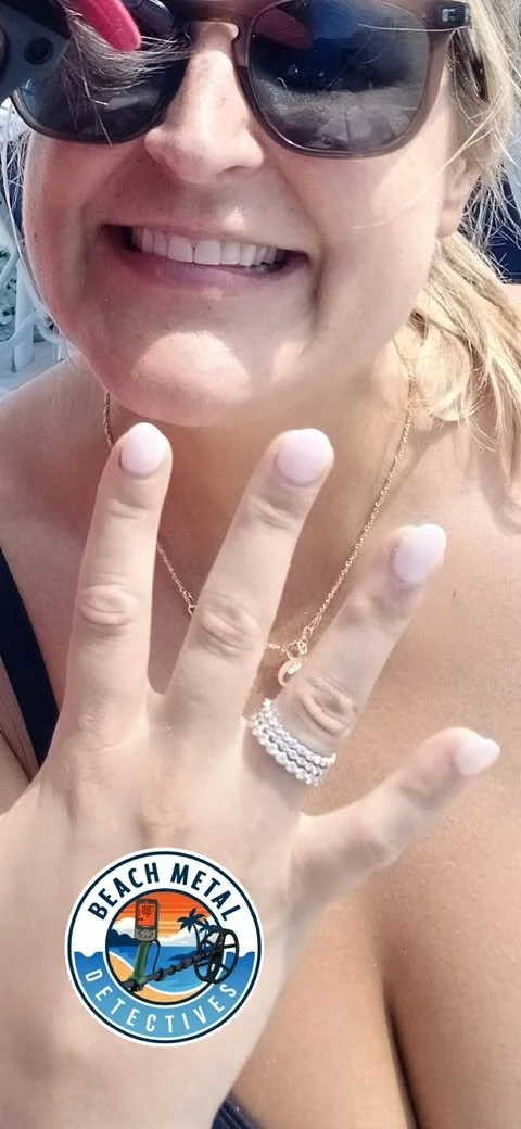 Smiling woman wearing sunglasses and jewelry, showing her hand with a stack of rings, with 'Beach Metal Detectives' sticker on her wrist.