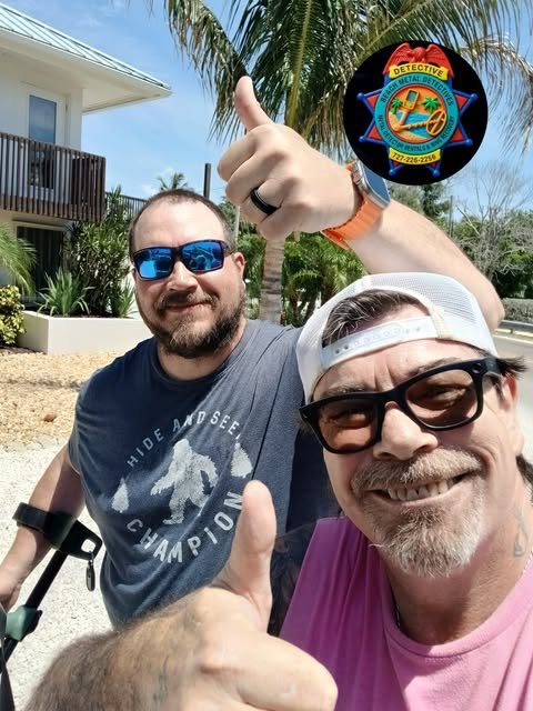 Two smiling men posing outdoors, one giving a thumbs up, with palm trees, a house, and a blue sky in the background. One man is wearing sunglasses and a beige cap, the other is wearing sunglasses and a gray T-shirt. The man in the pink shirt is holding a badge or patch that reads 'Detective.'