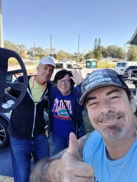 Three smiling people taking a selfie together outdoors, with a golf cart in the background. One person is giving a thumbs-up.