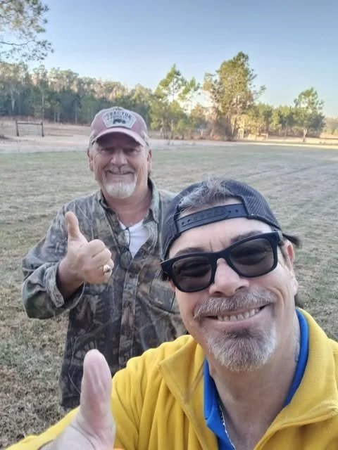 Two men smiling and giving a thumbs-up outdoors in a grassy area with trees in the background.