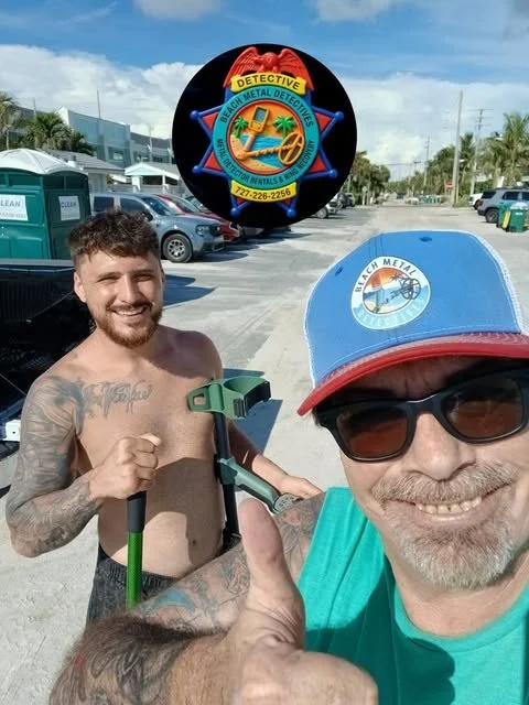 Two men at the beach, one smiling and wearing sunglasses and a blue cap, the other shirtless with tattoos, holding a metal detector, with parked cars and palm trees in the background.