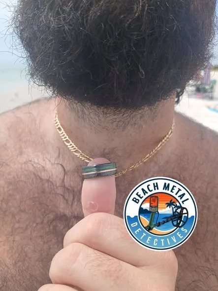 A man with a beard and chest hair holding a finger with a metal ring, showing a badge that reads 'Beach Metal Detectives' with a beach scene.