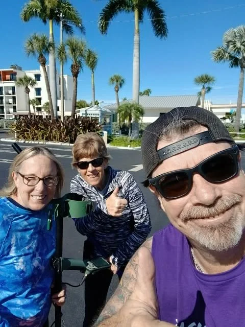 Three people taking a selfie outdoors on a sunny day with palm trees and buildings in the background. Two women and one man are smiling, wearing casual clothing and sunglasses.