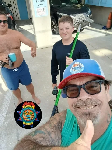 Three men smiling outdoors on a sunny day, one taking a selfie. The man in the foreground wears sunglasses, a red and blue hat, and a sleeveless shirt. The man in the middle holds a rake and a phone, wearing a dark shirt. The third man in the background is shirtless, wearing blue shorts, and holding a blue cup. There is a parking lot with a black car, a sign, and some orange cones.