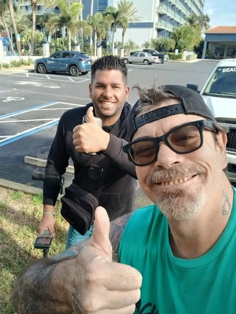 Two men are outdoors in a parking lot. One man in the foreground wears glasses, a backwards cap, and a teal shirt, and is giving a thumbs-up. The other man in the background has dark hair, a beard, and is smiling and holding a phone, giving a thumbs-up.