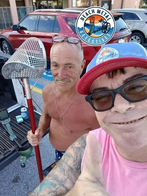 Two men taking a selfie in a parking lot, one shirtless holding a rake, the other wearing a pink shirt, red cap, and sunglasses, with a logo reading 'Beach Metal Detectives' in the background.