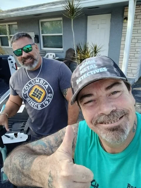 Two middle-aged men with beards smiling outdoors. One is taking a selfie, wearing a cap and a sleeveless teal shirt. The other is standing next to a pickup truck, wearing sunglasses and a navy T-shirt with a Columbia logo.