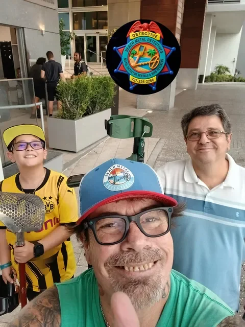 Three people taking a selfie outdoors in front of a building and a metal detector badge. The man in the front wears glasses, a green shirt, and a hat. The boy on the left wears glasses, a yellow sports jersey, and a yellow cap. The man on the right wears glasses and a white and blue polo shirt.