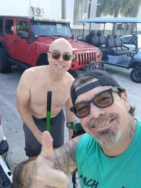 Two smiling men taking a selfie outdoors near a red jeep and golf carts. One man is shirtless and wearing sunglasses. The other wears a green shirt, sunglasses, and a backward cap, giving a thumbs-up.