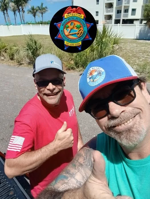 Two men taking a selfie outdoors, both wearing sunglasses and baseball caps, with a scenic background of palm trees and white buildings. One man in a red shirt and gray cap is giving a thumbs-up, while the other in a teal shirt and blue cap is smiling and giving a thumbs-up as well. A circular emblem for Beach Metal Detectors is floating above them.