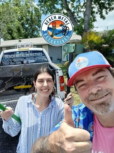 Two people taking a selfie in front of a Beach Metal Detectives vehicle, in a sunny outdoor setting with trees and a building, with a sign for Beach Metal Detectives above.