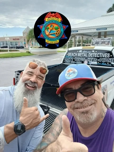Two men smiling and giving thumbs up in front of a pickup truck with a sign for a metal detector rental business. The truck has a window sign with contact info and the background shows a shopping area with stores and palm trees. A brightly colored circular logo for a metal detector service is above them.