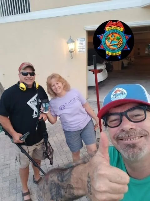 Three people posing outdoors near a building with a garage, smiling. The man on the right is giving a thumbs up, wearing glasses and a cap. The woman in the middle is holding a coffee cup. The man on the left is wearing headphones and holding equipment. A circular sign with a detection logo is above them.