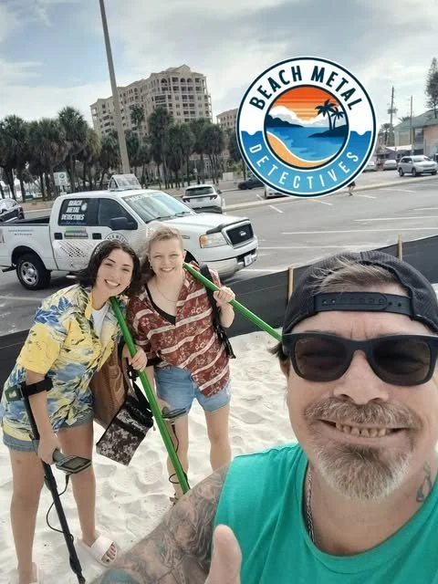 Three people, two young women and a man, are on the beach near parking lot, smiling for a selfie, with a Beach Metal Detectives logo in the top right corner, palm trees, parked cars, and buildings in the background.
