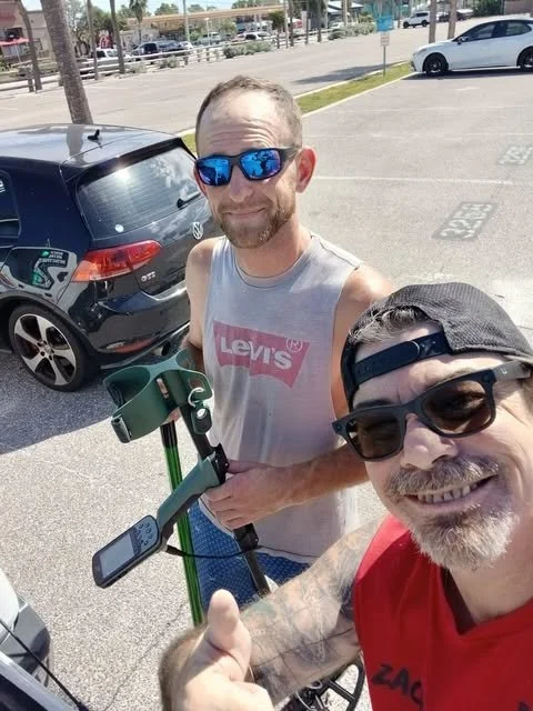 Two men taking a selfie in a parking lot with a scooter, wearing sunglasses, and smiling.