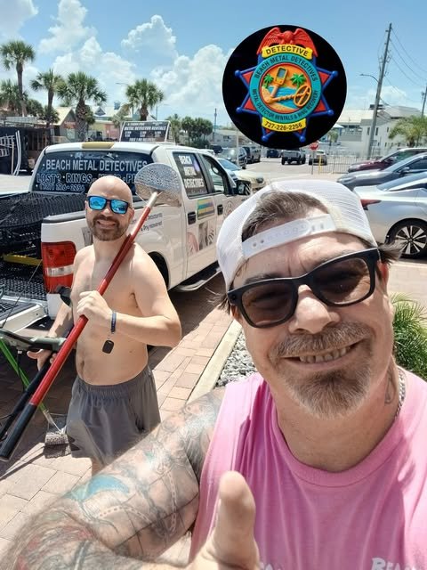 Two smiling men in sunglasses and casual clothing taking a selfie outdoors in a parking lot, with a beach metal detector service vehicle and palm trees in the background, under a partly cloudy sky.