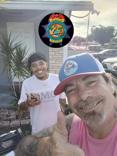 Two men smiling and taking a selfie outdoors. The man in the foreground wears a blue and red cap and a pink shirt, giving a thumbs-up. The man in the background wears a white shirt and holds a phone.