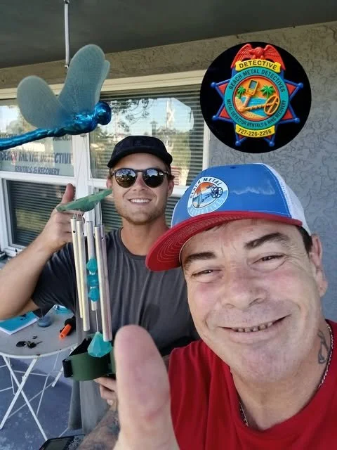 Two men taking a selfie outside, one wearing a red and blue cap and the other with sunglasses, holding a metal detector and a stuffed animal toy, with a weathered wall and a window in the background.