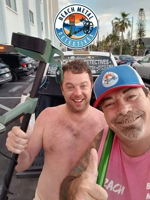 Two men taking a selfie in front of a beach-themed detective sign, with one man shirtless and holding a sledgehammer, and the other wearing a baseball cap and giving a thumbs up.