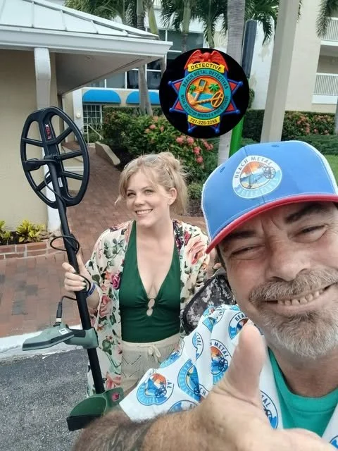 A man and a woman dressed as 1950s metal detector enthusiasts, with metal detectors in hand, outdoors in a tropical setting, smiling at the camera, man giving a thumbs up.