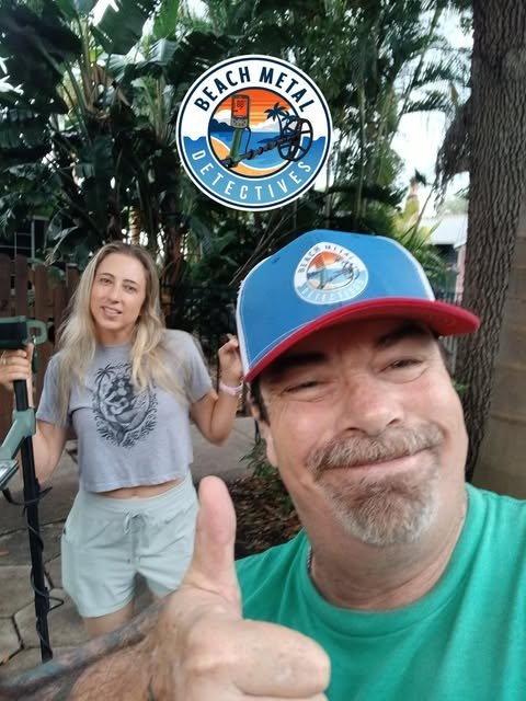 A man and woman outdoors, with a Beach Metal Detectives logo in the background. The man is wearing a blue and red cap and giving a thumbs up. The woman is holding a metal detector and wearing a gray t-shirt with a tiger design.