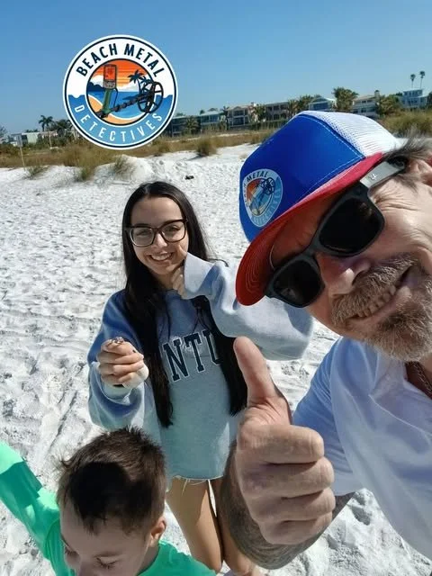 A man with sunglasses and a baseball cap giving a thumbs up, a woman with glasses smiling, and a child in a green shirt on a beach with sand, with a beach houses and a