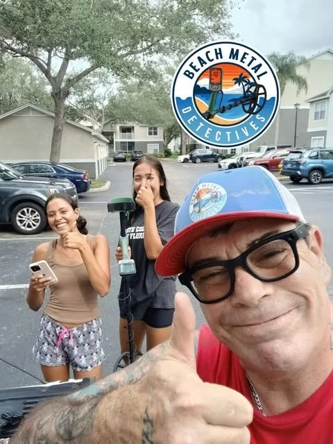 Three people taking a selfie outdoors in a parking lot with apartment buildings in the background, two young women and one man. The man is wearing glasses and a baseball cap, and giving a thumbs-up. One young woman is holding a phone, and the other girl is holding a rescue hook with the logo of Beach Metal Detectives.