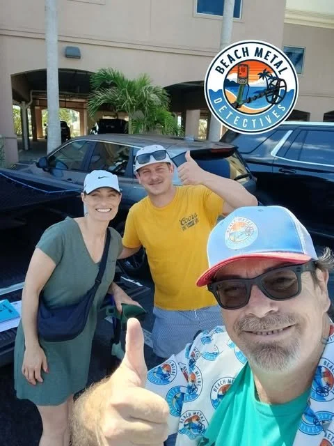 Three people smiling and taking a selfie outside, with a building, palm trees, and cars in the background. One person is wearing a cap and sunglasses, another is giving a thumbs-up, and the third is smiling. There is a "Beach Metal Detectives" logo in the upper right corner.