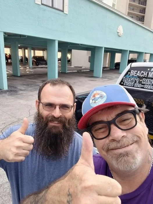 Two men smiling and giving thumbs up in a parking lot near a blue building; one man has a beard and glasses, the other wears glasses and a baseball cap.