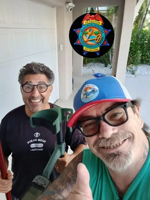 Two smiling men, one with glasses and a baseball cap, taking a selfie on a porch with a badge photo and some tools, with a background of palm trees and white gravel.