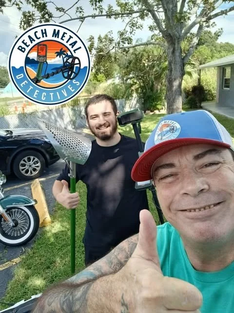 Two men take a selfie outdoors with a 'Beach Metal Detectives' logo in the top left corner. One man wears a blue cap with the same logo and gives a thumbs-up, while the other man holds a metal detector. They are in a grassy area near a tree, trees, and houses.