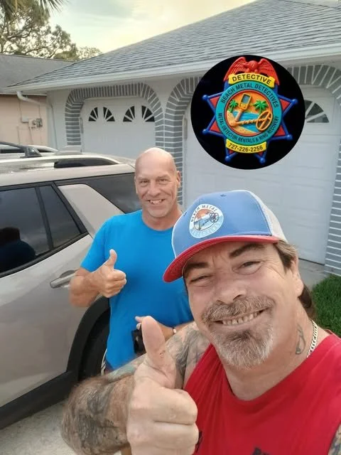 Two men taking a selfie in front of a house with a silver SUV parked nearby. The man in the foreground is wearing a red shirt and a blue and gray cap, giving a thumbs-up. The man in the background is wearing a blue shirt and also giving a thumbs-up. There is a circular logo or badge digitally added to the image with the words 'Detective Metal Detectors' surrounding a treasure chest and metal detector symbol.