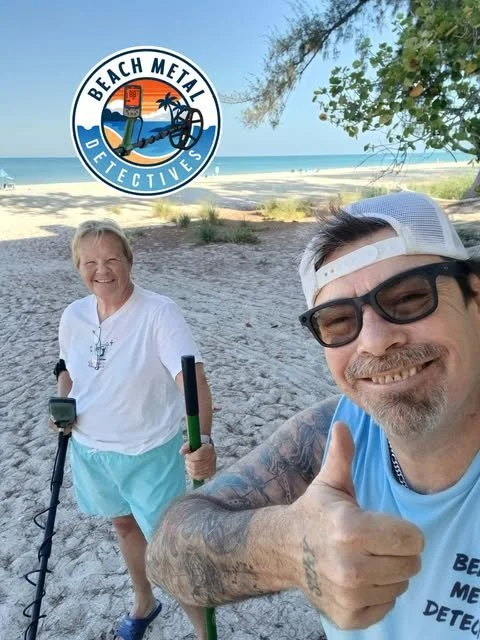 Two people on the beach, a woman with short blonde hair and a man wearing sunglasses and a white cap, taking a selfie. The woman is smiling and holding metal detectors. Behind them, there's a body of water and a clear sky. A badge with the logo of Beach Metal Detectives is in the top left corner.