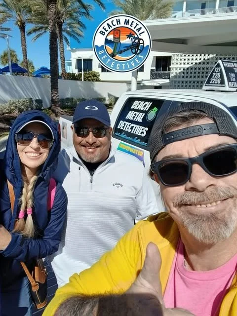 Three smiling people taking a selfie outdoors, with a car displaying a 'Beach Metal Detectives' sticker, palm trees, and a modern building in the background.