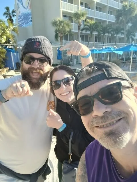Three people smiling and taking a selfie outdoors near a pool area with palm trees and a building in the background.