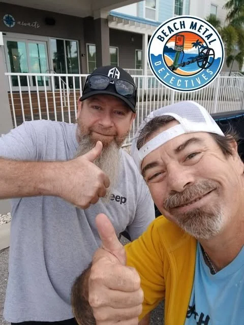 Two men smiling and giving thumbs up in front of a building, with a 'Beach Metal Detectives' logo in the top right corner.
