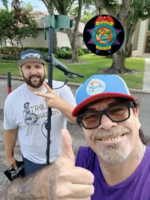 Two men outdoors, one smiling and giving a thumbs-up, the other smiling and making a peace sign. One man is wearing a cap and glasses with a badge, the other is wearing a white T-shirt and a hard hat with a badge. There is a large metal detector in the background and a badge with a patch showing a palm tree, sun, and a sheriff's badge.