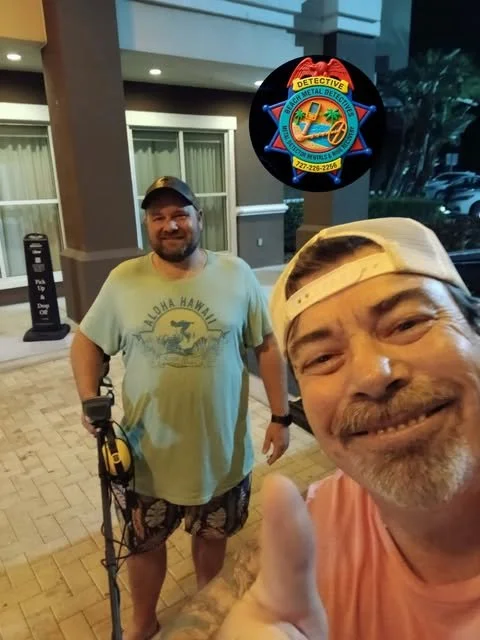 Two smiling men taking a selfie outdoors at night, one giving a thumbs-up. One man is wearing a light-colored cap, a pink T-shirt, and has a tattooed arm. The other man is wearing a baseball cap, a light green T-shirt with a graphic, and shorts. Behind them is a building with beige walls and large windows. A police-like badge sign reads 'Detective' with an emblem and contact information.