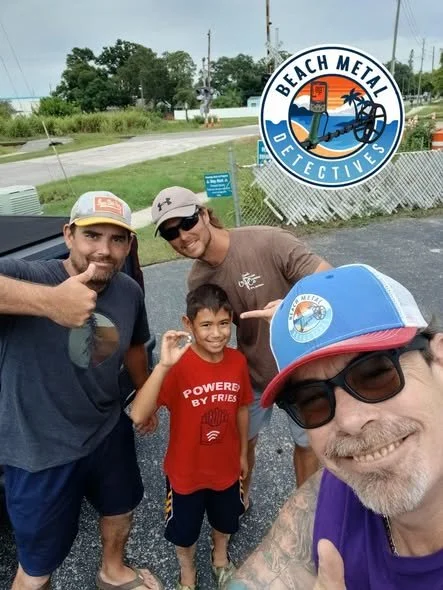 Four people, two adults and two children, are smiling and posing for a selfie outdoors during the daytime, with a sign for 'Beach Metal Detectives' visible in the background. They are casually dressed.